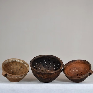 Op de afbeelding: Drie handgemaakte, rustieke, bruine aardewerken kommen met geperforeerde gaten. De kommen zijn in een rij gerangschikt, waarvan één gedeeltelijk open. De kommen hebben een natuurlijke, aardse esthetiek, wat wijst op een focus op functionaliteit en traditioneel vakmanschap.