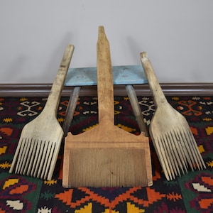 May include: Three antique wooden weaving tools, including two large combs and a central comb-like tool. The tools are light brown and rest on a colorful patterned rug. A small, light blue bench is in the background.