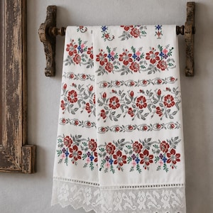 Puede incluir: Una toalla rectangular blanca con bandas horizontales de patrones florales en punto de cruz en tonos rojo, verde, azul y rosa. El borde inferior está rematado con un ancho y detallado encaje blanco con un diseño floral festoneado.