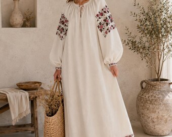 May include: A long-sleeved, off-white maxi dress featuring a gathered neckline with a tie closure and intricate floral embroidery in shades of red, burgundy, and green on the sleeves and hem, paired with a woven straw tote bag and brown sandals.