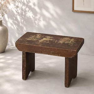 May include: A small, rectangular wooden stool with a dark brown finish and visible signs of wear, including patches of lighter, distressed wood on the top surface. The stool features four sturdy, block-style legs and a thick, solid seat.