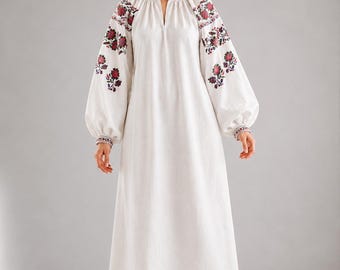 May include: A floor-length white dress featuring long, voluminous bishop sleeves with intricate red and dark floral embroidery. The garment includes a small V-neckline, gathered detailing at the shoulders, and a matching embroidered border at the hem.