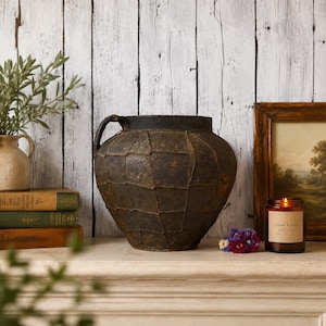 Op de afbeelding: Een grote, donkerbruine, getextureerde vaas met een handvat, op een witte schoorsteenmantel. De vaas heeft een uniek, patchwork-achtig ontwerp. Een brandende kaars in een glazen pot, boeken en een klein schilderij staan ook op de schoorsteenmantel.