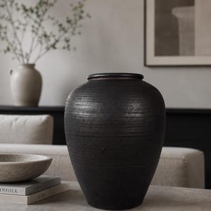 Può includere: Un grande vaso in ceramica color carbone scuro con una superficie strutturata e scanalata. Il vaso ha un corpo ampio e arrotondato e un bordo leggermente svasato. È un oggetto decorativo, adatto all'arredamento della casa.