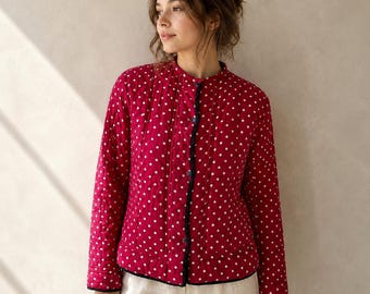 May include: A long-sleeved, quilted jacket in a vibrant red color featuring a repeating pattern of small white polka dots. The jacket has a round neckline, a front button closure with dark trim, and a straight hemline, worn over light-colored pants.