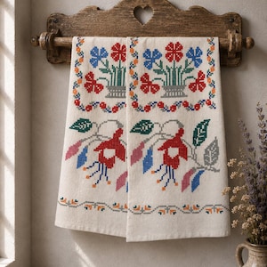 Puede incluir: Una toalla de lino color crema con un patrón de punto de cruz de flores azules y rojas en una cesta, junto con flores rojas tipo fucsia y hojas verdes. La toalla cuelga de un soporte de pared de madera marrón oscuro con un recorte de corazón.