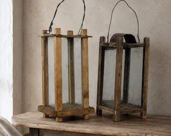 Può includere: Due lanterne rettangolari realizzate in legno chiaro e pannelli di vetro trasparente. Ogni lanterna presenta una base quadrata con quattro piccoli piedini e una sottile maniglia in filo metallico scuro fissata al telaio superiore per il trasporto.