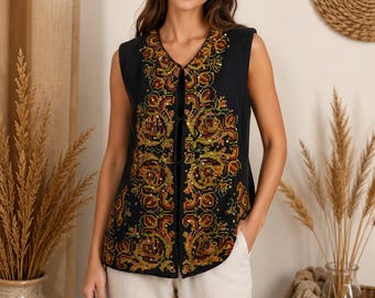 May include: A sleeveless black vest featuring an intricate, multi-colored embroidered pattern in shades of orange, yellow, and green, accented with small reflective sequins along the front panels and a simple hook-and-eye closure at the neckline.