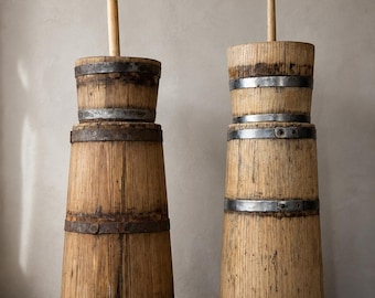 May include: Two tall, cylindrical wooden butter churns featuring vertical wood grain, weathered textures, and multiple dark, rusted metal bands encircling the bodies. Each churn includes a central wooden plunger handle extending from the top opening.