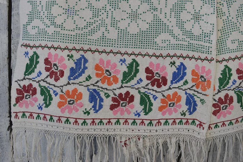 Puede incluir: Un tejido de color blanco roto que presenta un borde floral en punto de cruz con flores rosas, naranjas, rojo oscuro y azules intercaladas con hojas verdes. La tela incluye un patr&oacute;n geom&eacute;trico verde sobre las flores y flecos en el borde.