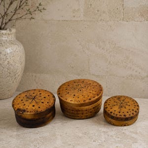 Puede incluir: Tres recipientes redondos de madera con tapa de diferentes tamaños, cada uno con un acabado natural marrón claro y patrones decorativos de color marrón oscuro grabados a mano con pequeñas flores, puntos y líneas geométricas.