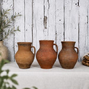 May include: Three antique terracotta jugs with handles, each with a unique weathered appearance. The jugs are displayed on a white surface against a rustic, white-painted wooden backdrop. The jugs vary in color from light brown to reddish-brown.