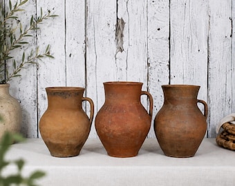 May include: Three antique terracotta jugs with handles, each with a unique weathered appearance. The jugs are displayed on a white surface against a rustic, white-painted wooden backdrop. The jugs vary in color from light brown to reddish-brown.