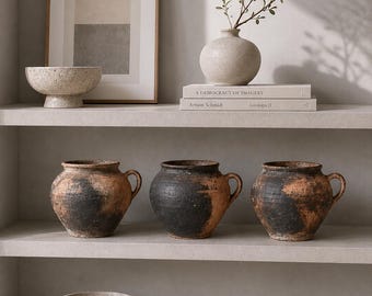 May include: A display of neutral-toned home decor items on shelves. Includes framed art, ceramic bowls, vases, and three rustic, handled pots with black and brown coloration. Books are stacked on the shelves.