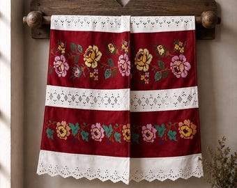 May include: A deep red kitchen towel featuring horizontal bands of colorful floral cross-stitch embroidery in yellow, pink, and green, interspersed with white eyelet lace trim and a scalloped bottom edge, hanging from a dark wooden wall rack.