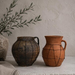 Può includere: Due brocche in terracotta con manici, ciascuna avvolta in una sottile rete metallica scura. La brocca a sinistra è marrone scuro con una superficie ruvida e usurata, mentre quella a destra è di colore arancione terracotta con finitura liscia.