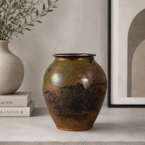 May include: A handcrafted, round ceramic vase with a wide base and a narrow opening. The vase features a blend of earthy tones, including green, brown, and black, creating a rustic aesthetic. The vase is approximately 12 inches tall.