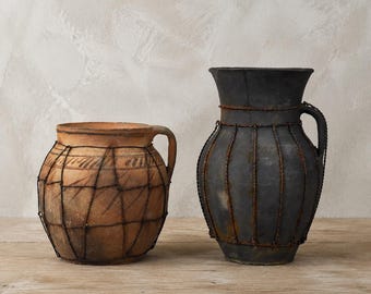 May include: A short, round terracotta pot with a handle and a tall, dark gray ceramic pitcher with a handle. Both vessels are wrapped in a decorative, thin, rusted metal wire cage that creates a grid pattern around the exterior of each piece.