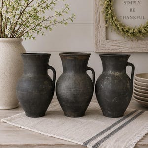 Può includere: Tre brocche in ceramica grigio antracite scuro con una finitura opaca e usurata. Ogni brocca presenta un corpo arrotondato, un bordo svasato e un unico manico laterale. I recipienti sono disposti in fila, mostrando una consistenza rustica uniforme.