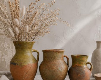 May include: Three rustic ceramic pitchers with textured, weathered surfaces in shades of olive green, brown, and terracotta. The tallest pitcher on the left contains a large arrangement of dried wheat stalks and fluffy white grasses.