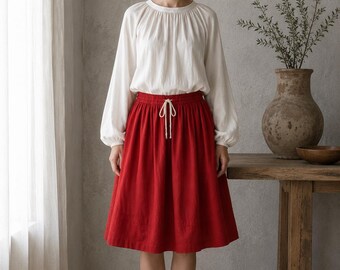 May include: A white long-sleeved blouse with a gathered round neckline and cuffed sleeves paired with a knee-length red skirt featuring an elasticized waistband and a central drawstring tie. The skirt has a simple, straight-cut silhouette.