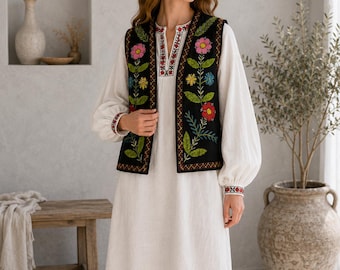 May include: A white knee-length dress with long, full sleeves and a black vest featuring colorful floral embroidery. The vest has a V-neckline with intricate red and white patterns, while the sleeve cuffs also display matching red and white embroidery.
