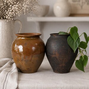 Può includere: Due vasi in terracotta con corpi arrotondati e bordi larghi. Il vaso a sinistra presenta una finitura smaltata color ambra lucido, mentre il vaso a destra ha una texture opaca color nero carbone con segni visibili di usura e invecchiamento.