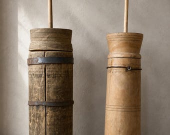 May include: Two tall, cylindrical wooden butter churns with vertical plungers. The left churn features weathered wood with dark metal bands, while the right churn is made of lighter, smoother wood secured with thin wire rings.