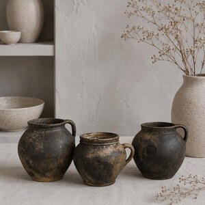 May include: Three dark brown, weathered ceramic pots with rounded bodies and single side handles. Each vessel features a textured, mottled surface with visible signs of age and wear, arranged in a row on a light-colored fabric surface.