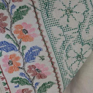 Puede incluir: Un tejido de color crema con un intrincado bordado de punto de cruz. El dise&ntilde;o muestra un patr&oacute;n floral con flores rosas, naranjas y azules junto a hojas verdes. Un borde geom&eacute;trico verde enmarca el lateral y flecos blancos cuelgan de un lado.