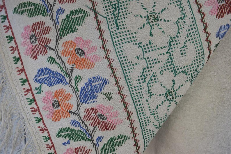 Puede incluir: Un tejido de color crema con un intrincado bordado de punto de cruz. El dise&ntilde;o muestra un patr&oacute;n floral con flores rosas, naranjas y azules junto a hojas verdes. Un borde geom&eacute;trico verde enmarca el lateral y flecos blancos cuelgan de un lado.