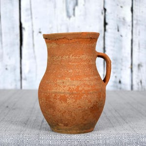Puede incluir: Jarra rústica de color terracota con asa. La jarra de barro tiene una superficie texturizada y un borde ligeramente irregular. El fondo es una pared de madera blanca y un mantel gris.