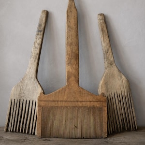 May include: Three wooden flax combs with long handles and fine, vertical teeth. The central comb features a wider, rectangular base with engraved horizontal lines, while the two flanking combs have narrower, tapered heads and weathered wood grain.
