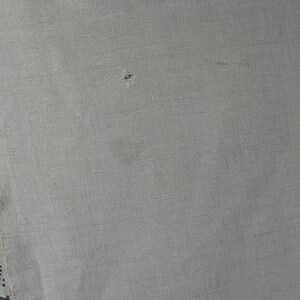 Puede incluir: Un trozo de tela tejida de color beige claro con un borde vertical de bordado en punto de cruz. El bordado muestra un patr&oacute;n repetitivo en tonos verde, rojo y rosa. La tela tiene una textura visible y un peque&ntilde;o agujero cerca del centro.
