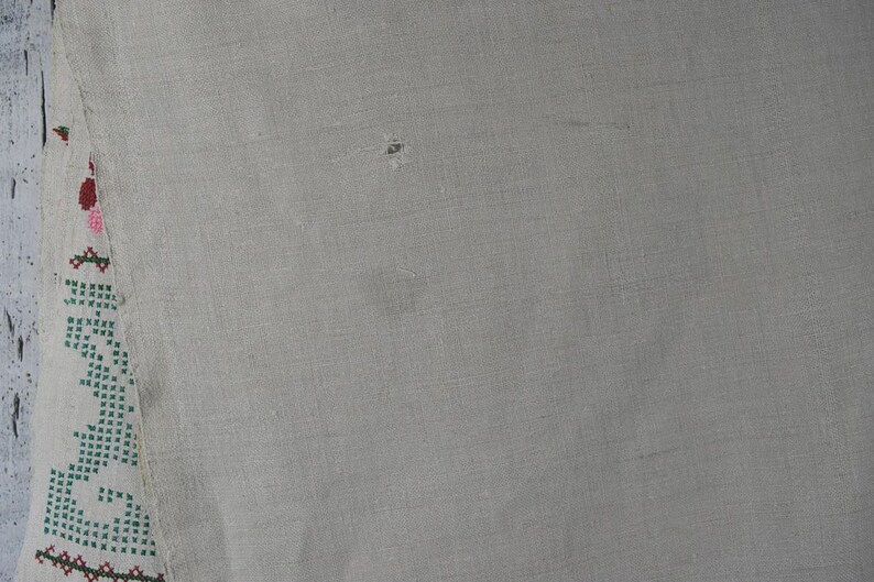 Puede incluir: Un trozo de tela tejida de color beige claro con un borde vertical de bordado en punto de cruz. El bordado muestra un patr&oacute;n repetitivo en tonos verde, rojo y rosa. La tela tiene una textura visible y un peque&ntilde;o agujero cerca del centro.