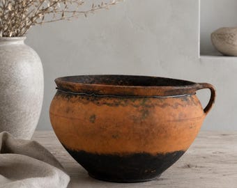 May include: A round terracotta bowl with a wide opening and a single side handle. The exterior features a two-tone finish with a warm orange-brown upper section and a dark, charred black base, accented by a thin wire wrapped around the rim.
