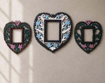 May include: Three heart-shaped picture frames with intricate cut-out details. The two outer frames are dark green with pink heart and flower accents. The central, larger frame is black and white with light blue floral accents and a black center.