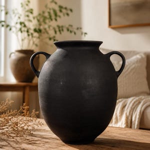 May include: A large, black, matte-finish vase with two loop handles. The vase has a rounded body and a slightly flared rim. It is sitting on a wooden table. A smaller vase and greenery are in the background.