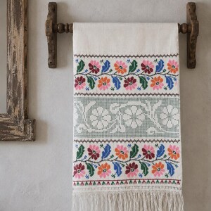 Puede incluir: Un tejido rectangular de color blanco roto colgado de una barra de madera, con bandas horizontales de motivos florales en punto de cruz en rosa, naranja, azul y verde, un panel central decorativo en verde salvia y flecos en el borde inferior.