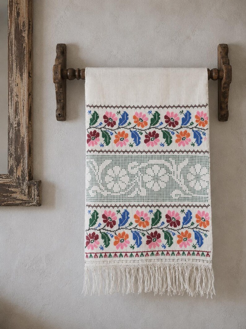 Puede incluir: Un tejido rectangular de color blanco roto colgado de una barra de madera, con bandas horizontales de motivos florales en punto de cruz en rosa, naranja, azul y verde, un panel central decorativo en verde salvia y flecos en el borde inferior.