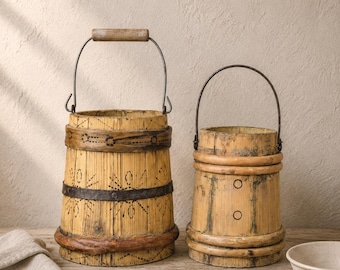 May include: Two cylindrical wooden buckets with metal handles and wooden grips. The larger bucket features dark painted floral patterns and a metal band, while the smaller bucket has two horizontal wooden rings and two circular markings.