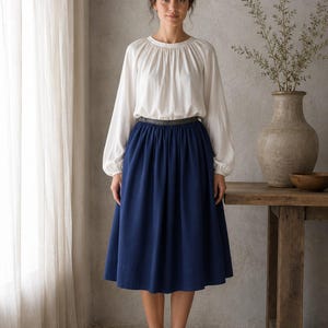 May include: A long-sleeved white blouse with a gathered round neckline and a dark blue midi skirt featuring a wide waistband. The skirt has a full, pleated silhouette that falls below the knees, paired with simple beige flat shoes.