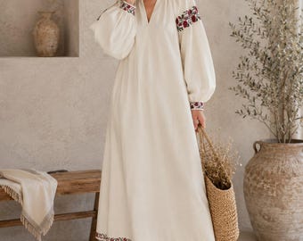 May include: A cream-colored maxi dress featuring a V-neckline, long billowy sleeves, and a hemline finished with intricate red and black floral embroidery. The embroidery also accents the shoulders and cuffs of the garment.