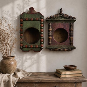 May include: Two rectangular wooden wall-mounted display cases with circular openings. The left case is green with red beaded trim and a cross top. The right case is dark red with green architectural molding and a finial top. Both have hollow centers.