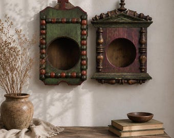May include: Two rectangular wooden wall-mounted display cases with circular openings. The left case is green with red beaded trim and a cross top. The right case is dark red with green architectural molding and a finial top. Both have hollow centers.