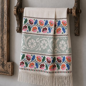 Puede incluir: Un tejido rectangular de color crema con un intrincado bordado de punto de cruz, que presenta bandas horizontales de coloridos motivos florales en tonos rosa, naranja, azul y verde, rematado con un largo y delicado fleco blanco en el borde inferior.
