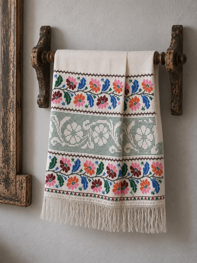 Puede incluir: Un tejido rectangular de color crema con un intrincado bordado de punto de cruz, que presenta bandas horizontales de coloridos motivos florales en tonos rosa, naranja, azul y verde, rematado con un largo y delicado fleco blanco en el borde inferior.