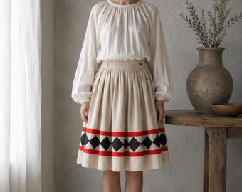 May include: A cream-colored long-sleeve blouse with a gathered neckline and a beige knee-length skirt featuring a horizontal band of black diamonds bordered by two thin red stripes. The skirt has a fitted waistband with a single visible button.