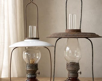 May include: Two vintage-style oil lamps with amber glass bases and clear glass chimneys. The left lamp features a white enamel shade, while the right lamp has a larger, rusted metal shade. Both include metal wire frames and top hanging loops.