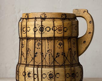 May include: A cylindrical wooden mug featuring a light brown finish with dark brown pyrography patterns of circles, crosses, and floral motifs. The mug is reinforced with vertical and horizontal dark brown twine and has a curved wooden handle.